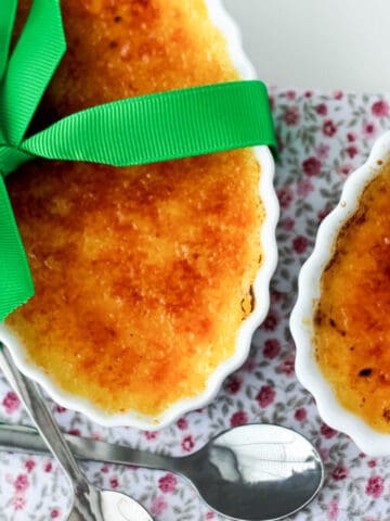 2 French Crèmes brûlées in a white dish with a green ribbon in a bow and 2 spoons on the side