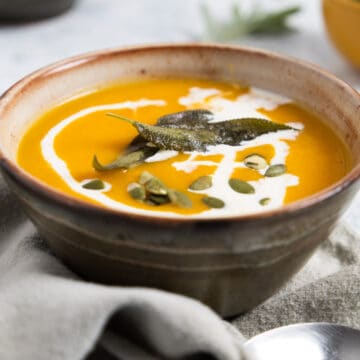 brown bowl with orange soup in it with a drizzle of cream and sage leaves on top.