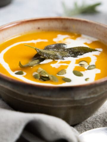 brown bowl with orange soup in it with a drizzle of cream and sage leaves on top.