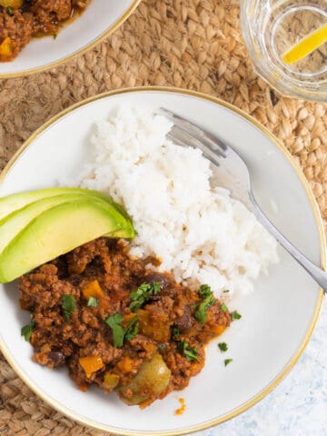 Ground beef served in white plate wit some white rice and 3 slices of avocados, there are 2 glasses of water with slices of lemon.