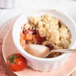 apple and strawberry crumble half eaten in a white ramekin and a fresh strawberry on a pink plate.