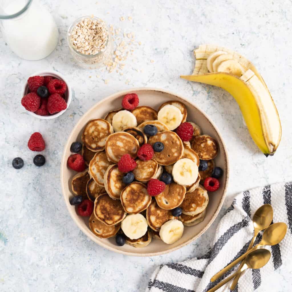 shallow plate filled with mini pancakes and fruits and a banana sliced on the side, oats, raspberries in a ramekin.