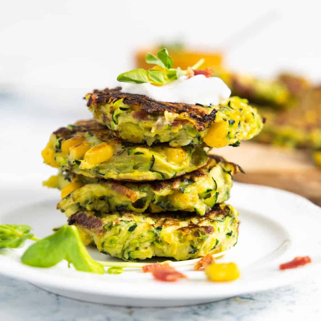 stack of 4 zucchini, corn and bacon fritters on a white plate with a dolop of sour cream on top.