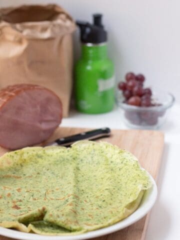 Side picture of savory crepes, green with grapes in the back and some ham.