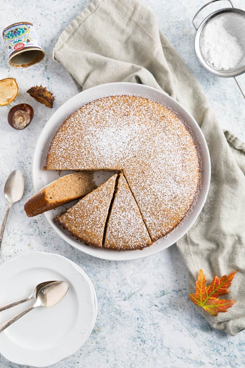 Top view of the chestnut cake with 3 slices already cut and one slice is flipped. A tea towel on the side with a red leaf and the empty chestnut container.