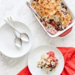 Oven dish filled with a berry crumble. One share is served on a white plate and another plate on the side with 2 spoons.