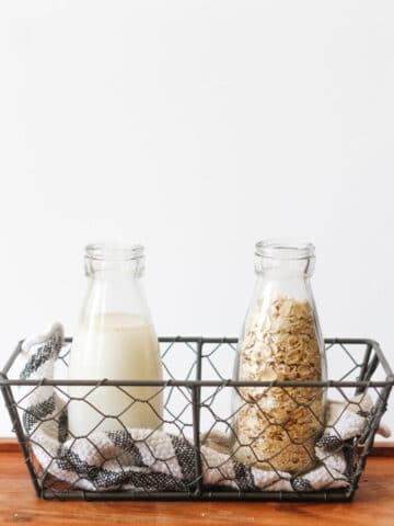 Preparation of homemade oat milk: 2 small bottles in a mesh basket with a black and white dish towel. One bottle is filled with oat milk and one with oats