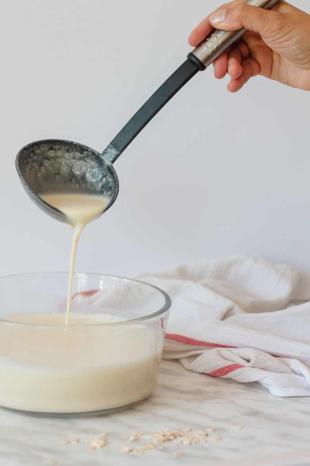 Oat milk French crêpes batter in a clear bowl with a black ladle pouring some, white dish towel on the side.