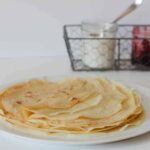 Stack of Oat Milk French crêpes on a white plate with a sugar and jam in the background