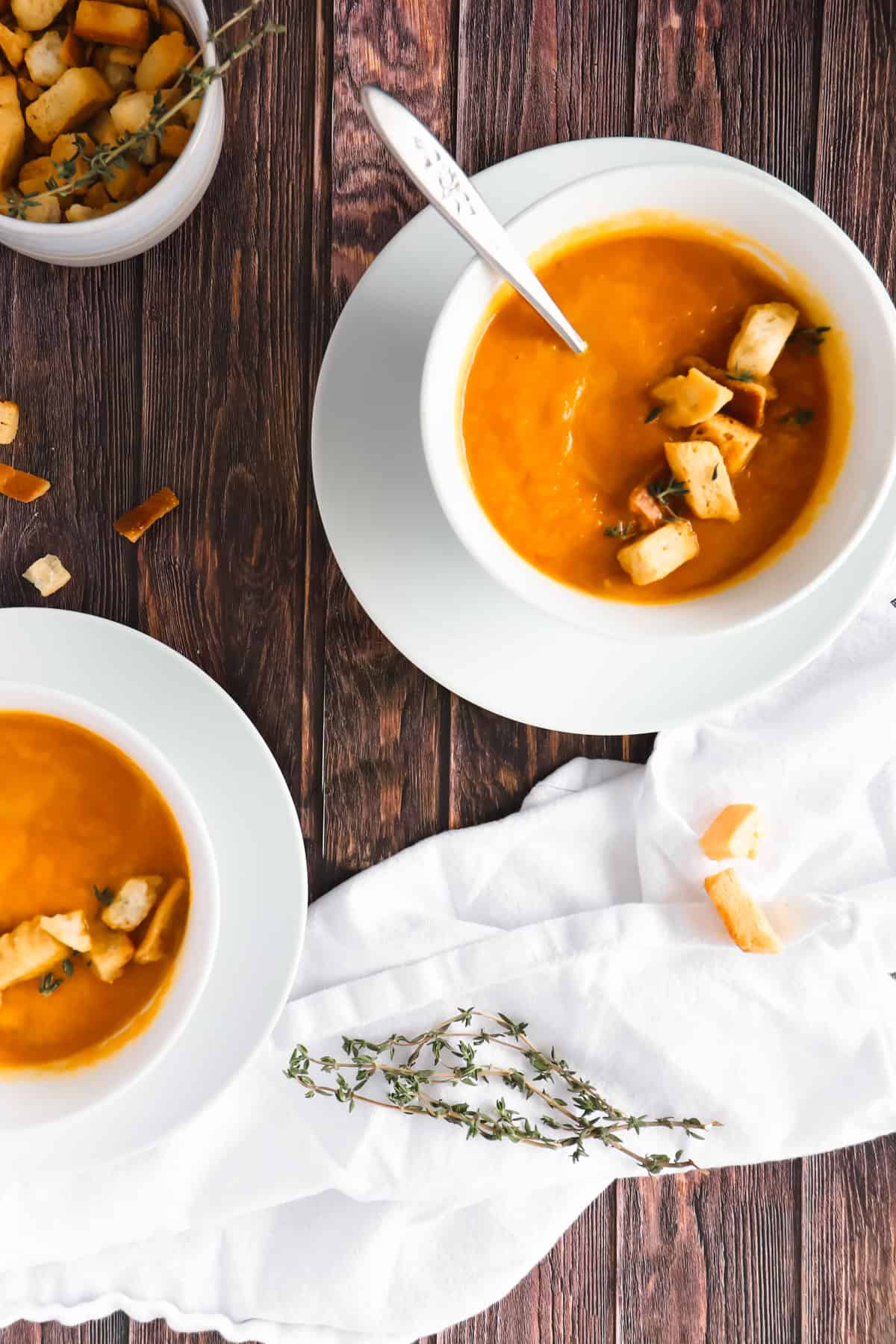 two white bowls on white plates with carrots soup in it and some croutons on top. 