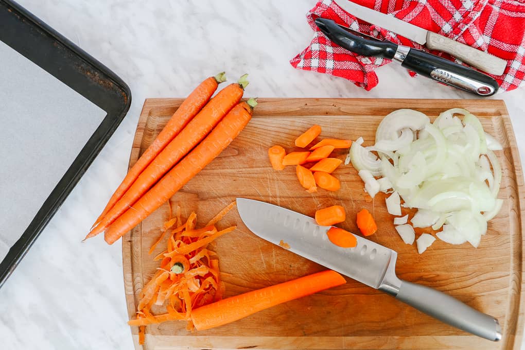 Top view of a wooden chopping board with a knife and some carrots chopped and chopped onion on the side.