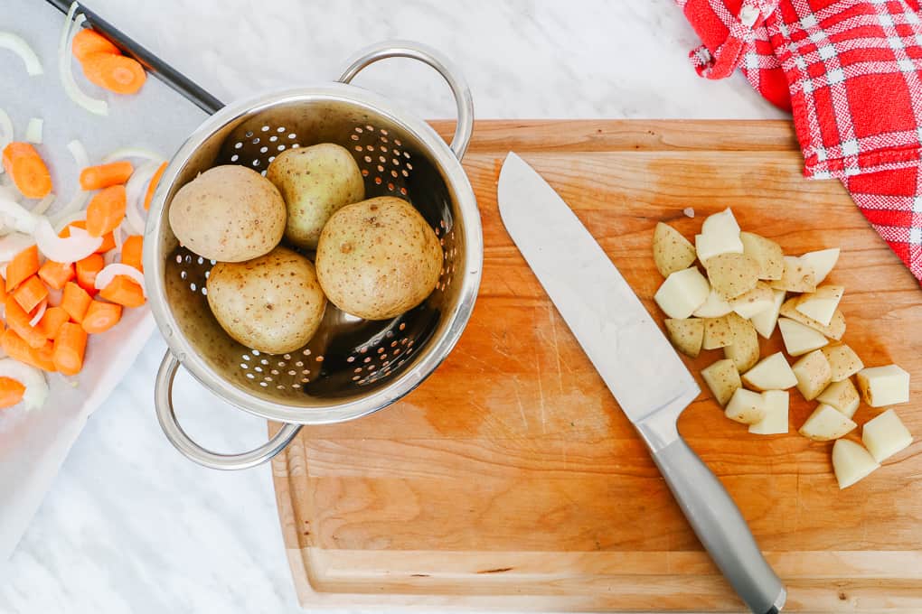 Top view of a wooden chopping board with a knife and some potatoes cubed and some in the metal colandor.