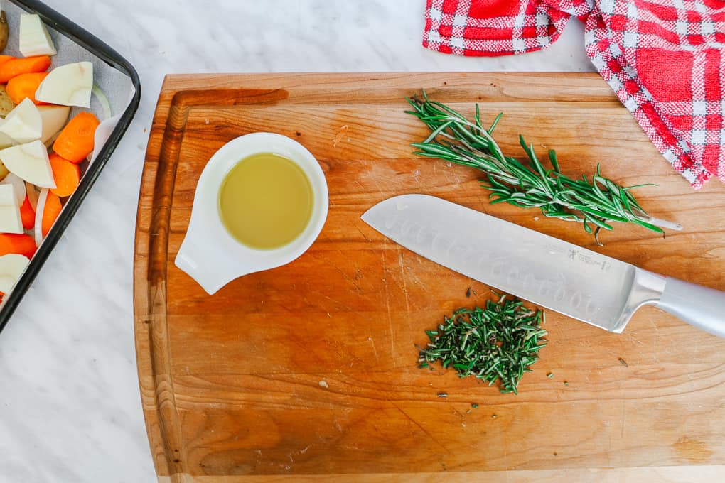 Top view of a wooden chopping board with a knife with chopped garlic, olive oil and rosemary.