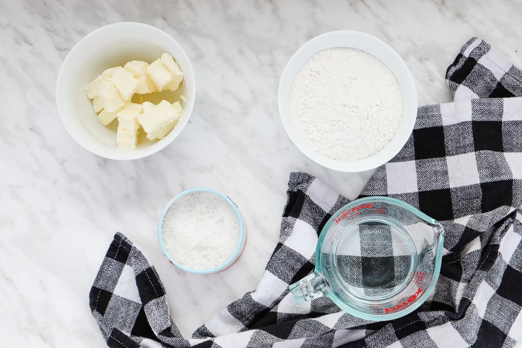 top view of ingredients for a pastry crust, butter, flour, salt and water and a black and white tea towel on the side