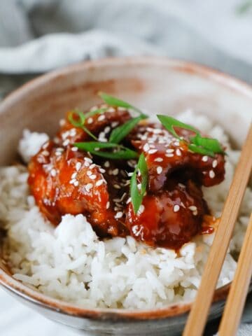 Close side view of 2 brown bowls with sweet and sour chicken served over white rice, some chop sticks on top
