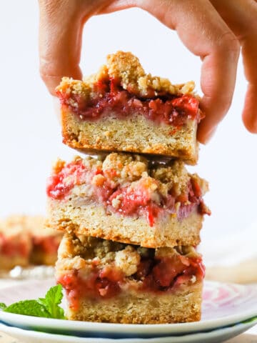3 strawberry crumble bars stacked on 2 small plates on top of a vintage book. 2 fresh strawberries on the side.