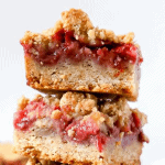 3 strawberry crumble bars stacked on 2 small plates on top of a vintage book. 2 fresh strawberries on the side.