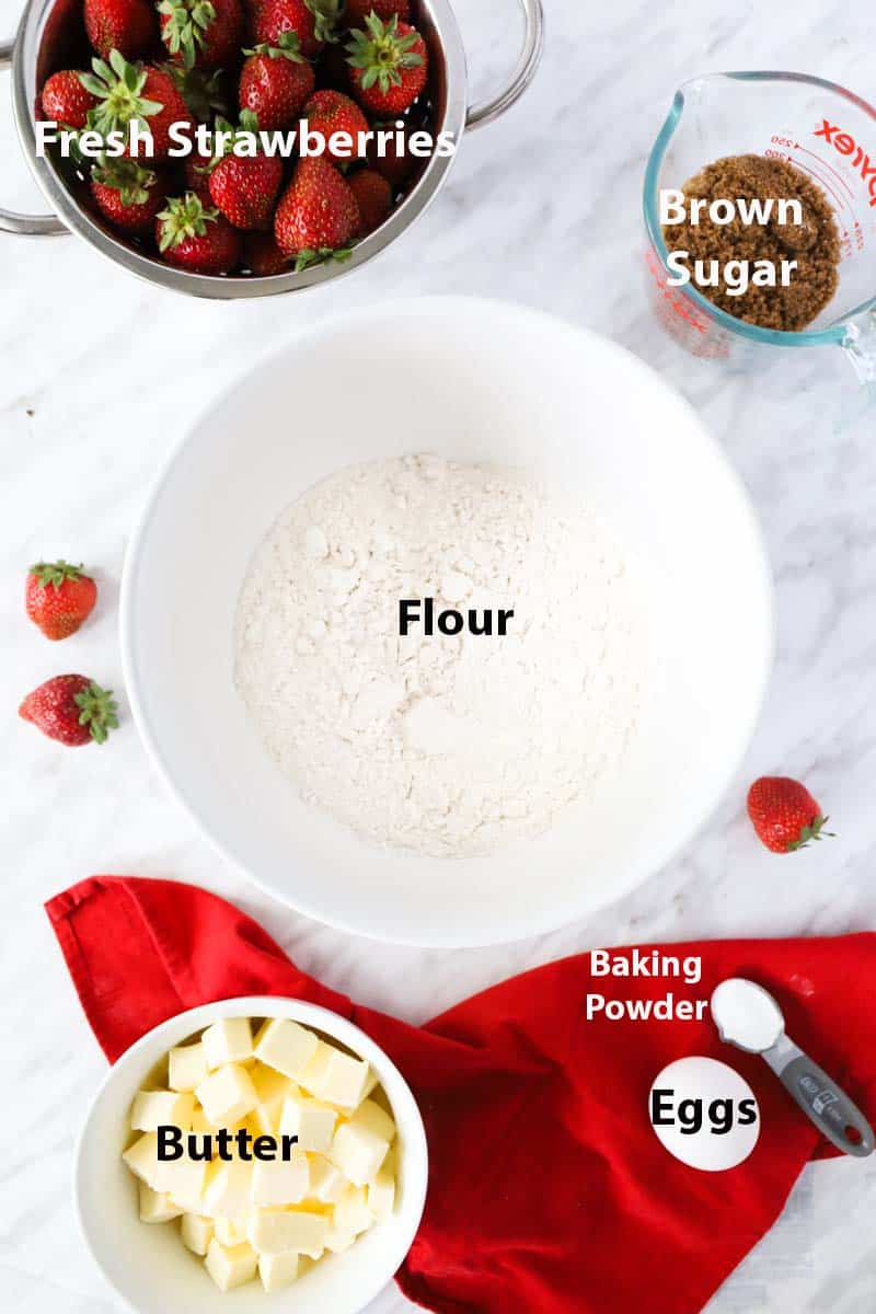 Top view of a white bowl with flour, small bowl with cubes of butter, colander of fresh strawberries. Red tablecloth on the side.