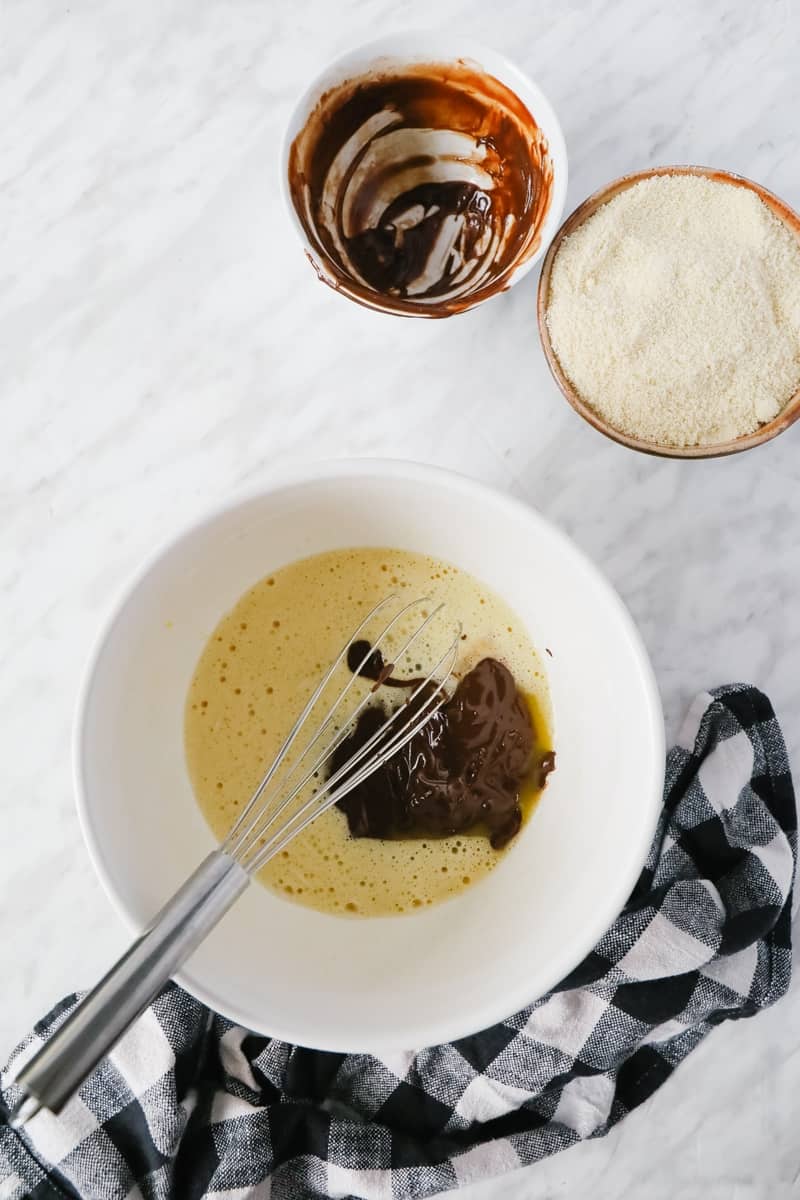 step of a chocolate cake: melted chocolate in a white bowl with beaten eggs and a bowl of almond flour on the side. 