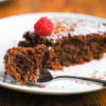 close up of a piece of chocolate almond cake in a spoon from a slice on a white plate with a raspberry on the side