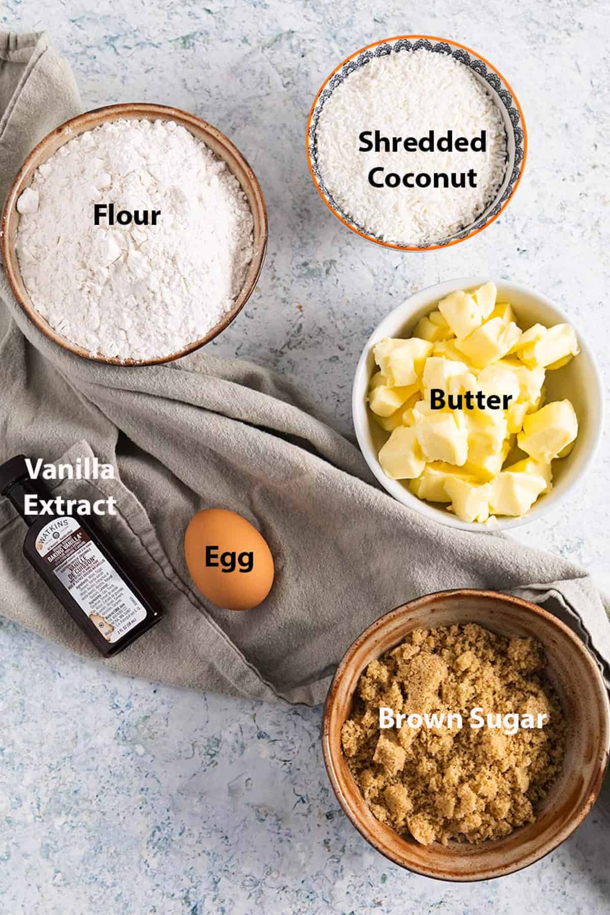 top view of ingredients to make coconut sugar cookies: flour, shredded coconut, butter, egg, vanilla extract and brown sugar.