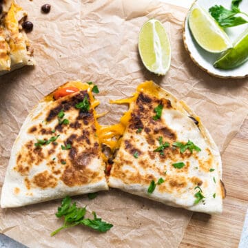 top view of some quesadillas on a brown paper. The quesadilla is sliced and the cheese is pulled. there is some cilantro in a small ramekin and slice of lime