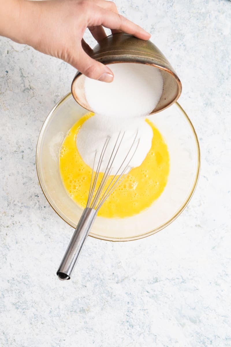 top view of a transparent mixing bowl with beaten eggs and sugar being poured