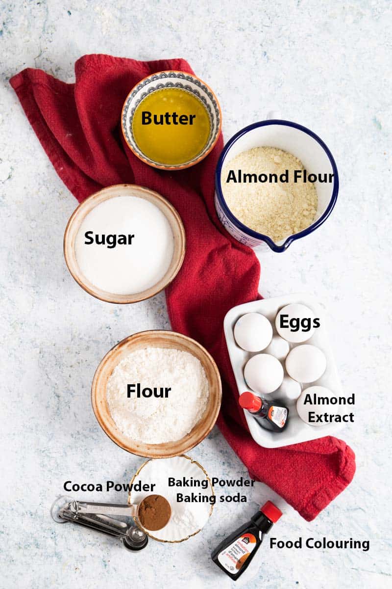 Top view of ingredients in ramekins: melted butter, almond flour, sugar, flour, eggs, almond extract, baking powder and baking soda, food colouring and a red tea towel