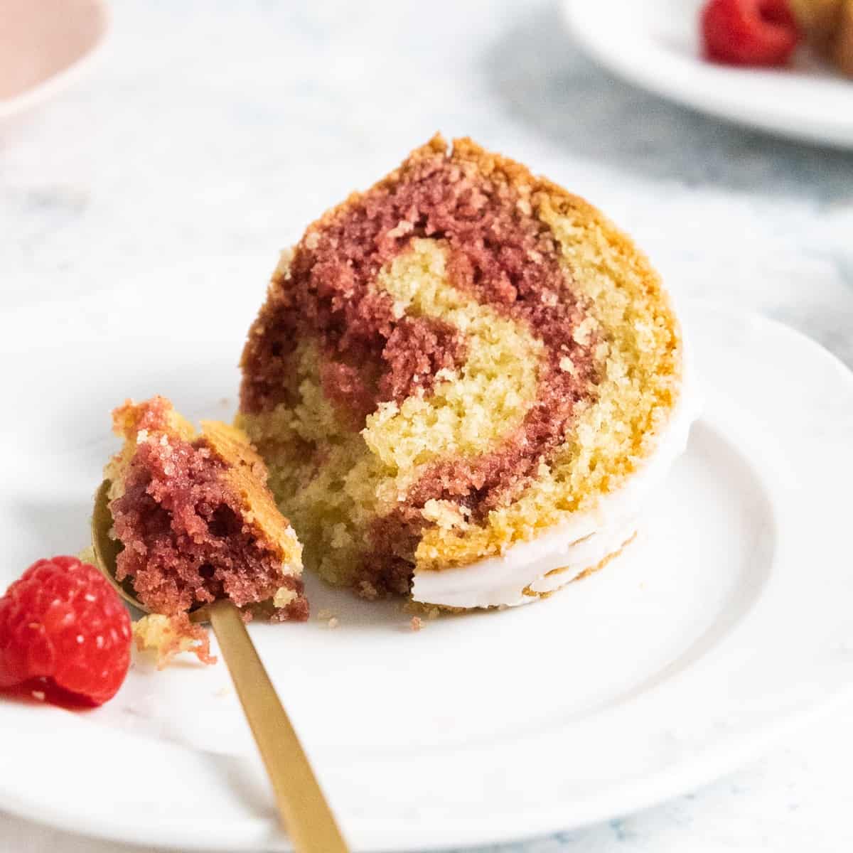 close up of a slice of Red Velvet Almond Marble Bundt Cake in a white plate with a golden spoon and raspberry on the side. 