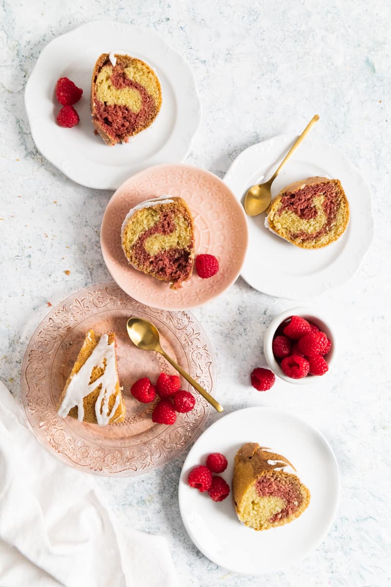 Top view of 5 plates with slices of Red Velvet Almond Marble Bundt Cake in pink plates and white ones with golden spoons and a ramekin of raspberries on the side