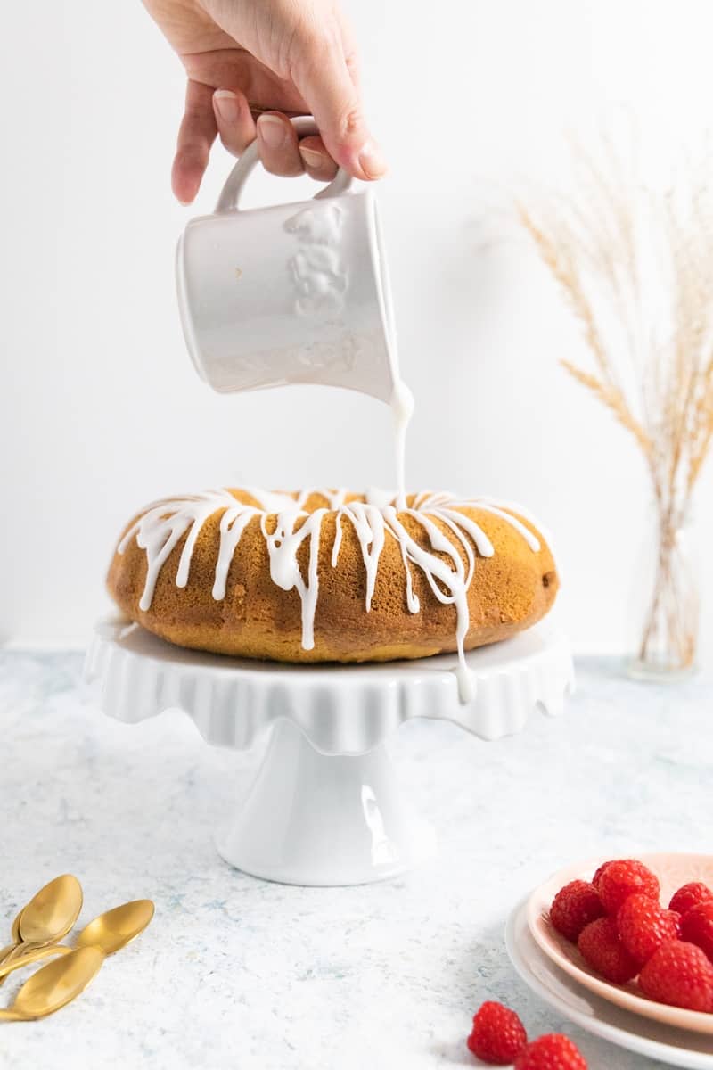 Side view of a bundt cake on a white cake stand and a hand drizzling some white icing. Fresh raspberry on the front right of the picture