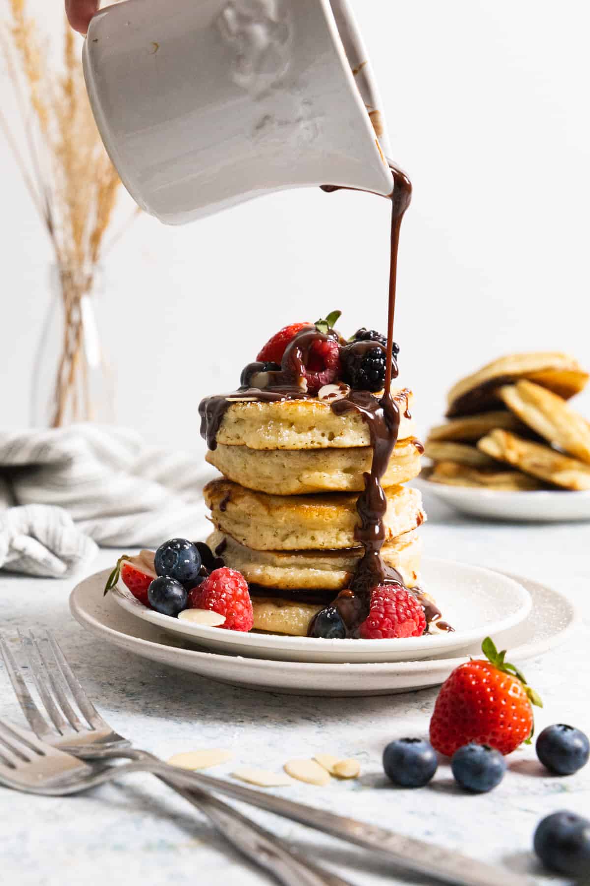 Stack of almond milk pancakes with a strawberries, raspberries, blackberries and chocolate being poured on top