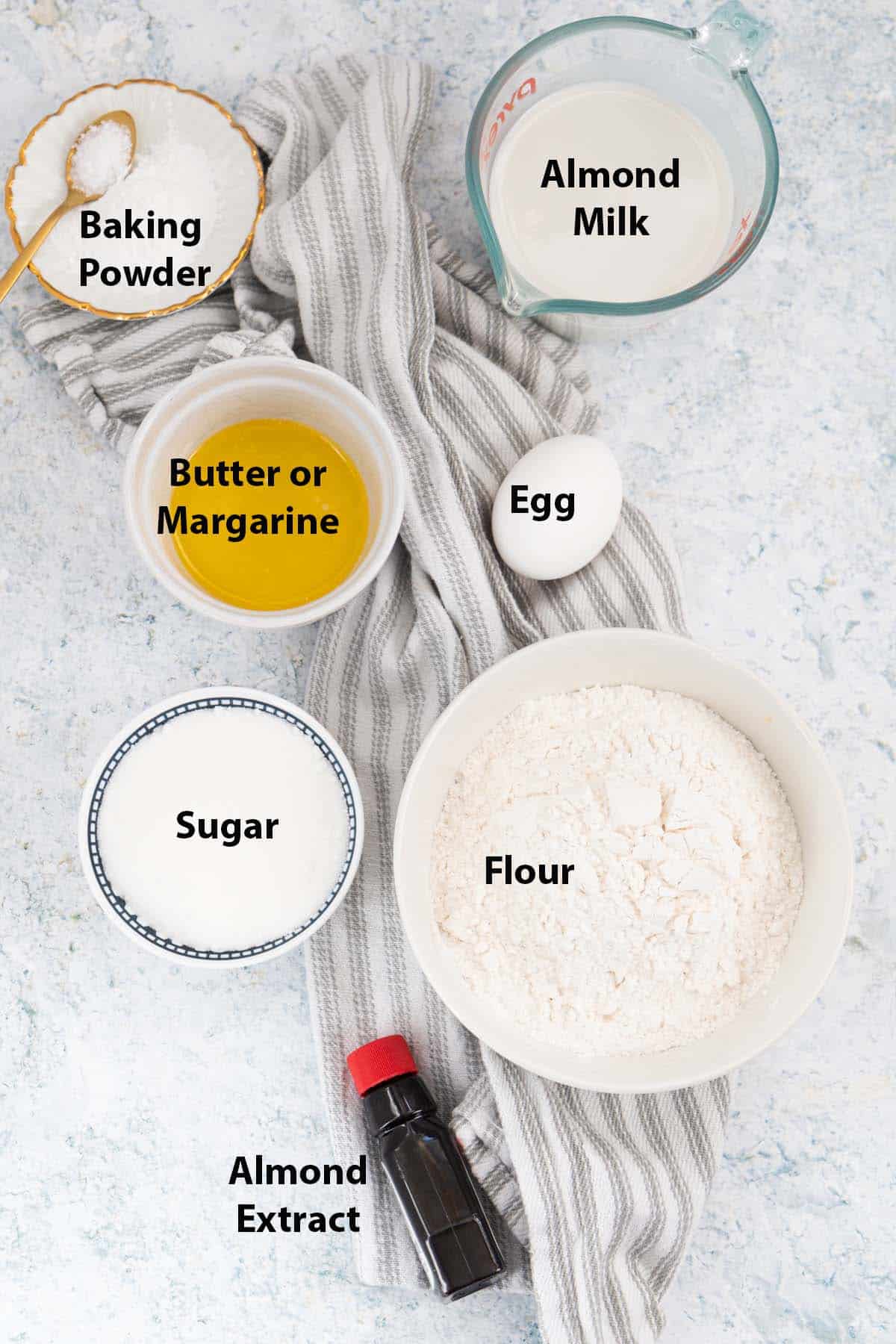 Top view of labelled ingredients in small bowls and ramekins: sugar, butter, flour, egg, baking powder and almond milk