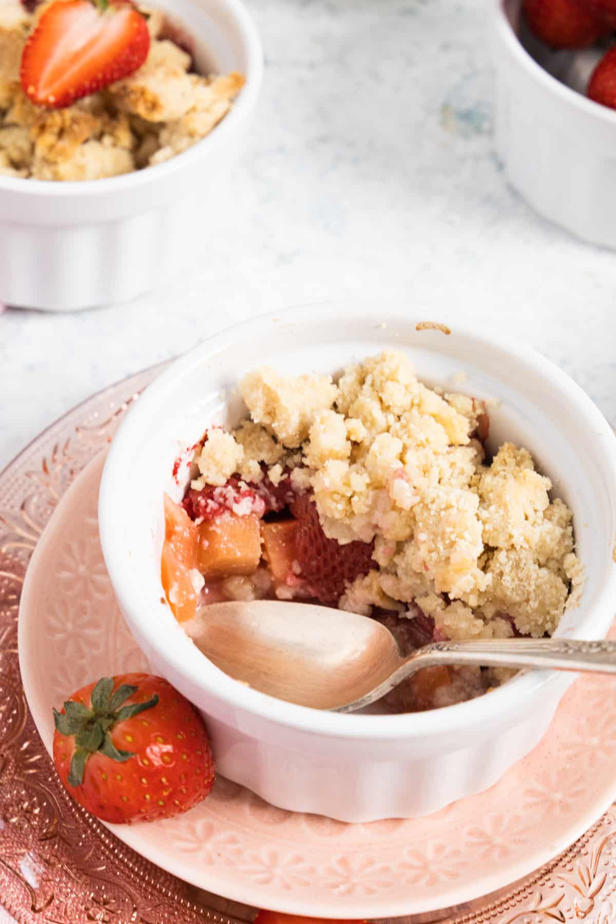 apple and strawberry crumble half eaten in a white ramekin and a fresh strawberry on a pink plate. 