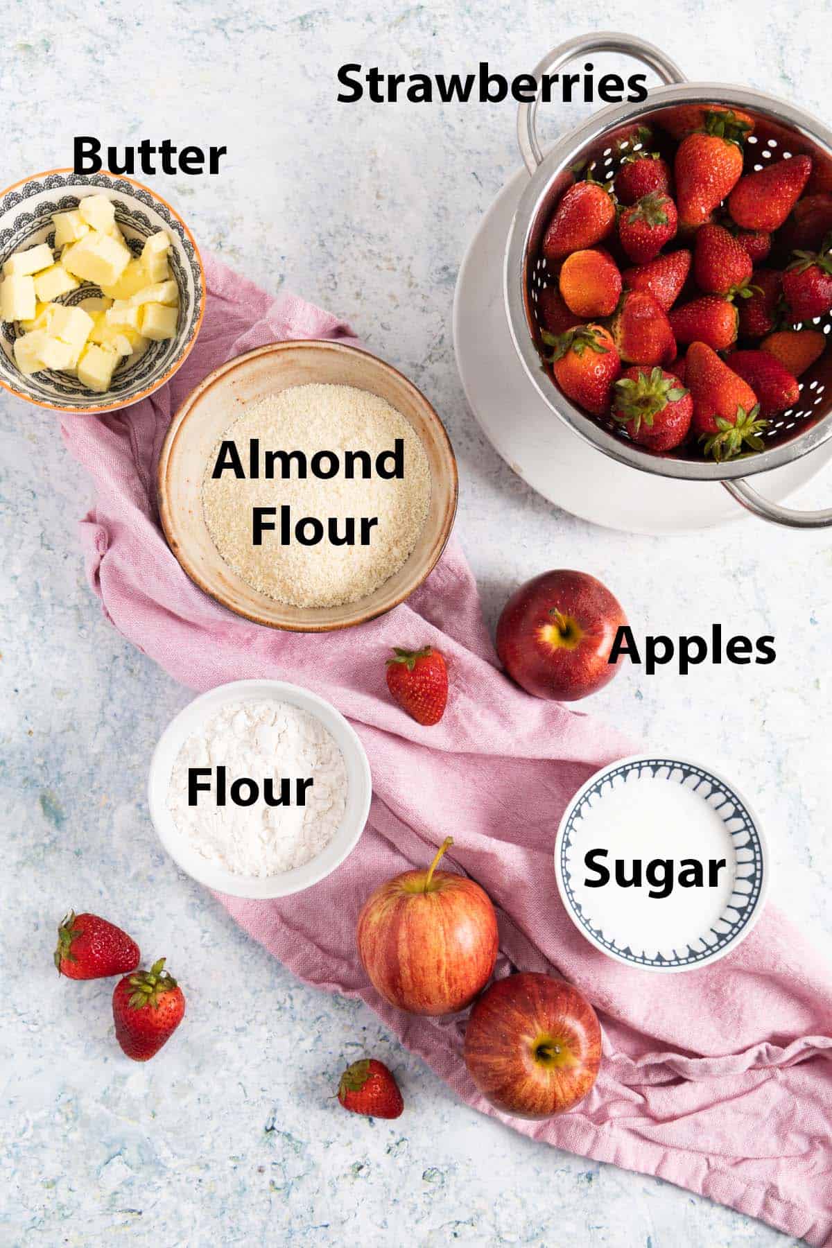 all the ingredients to make apple and strawberry crumble laid out and labelled: butter, strawberry, almond flour, apples, flour, sugar.
