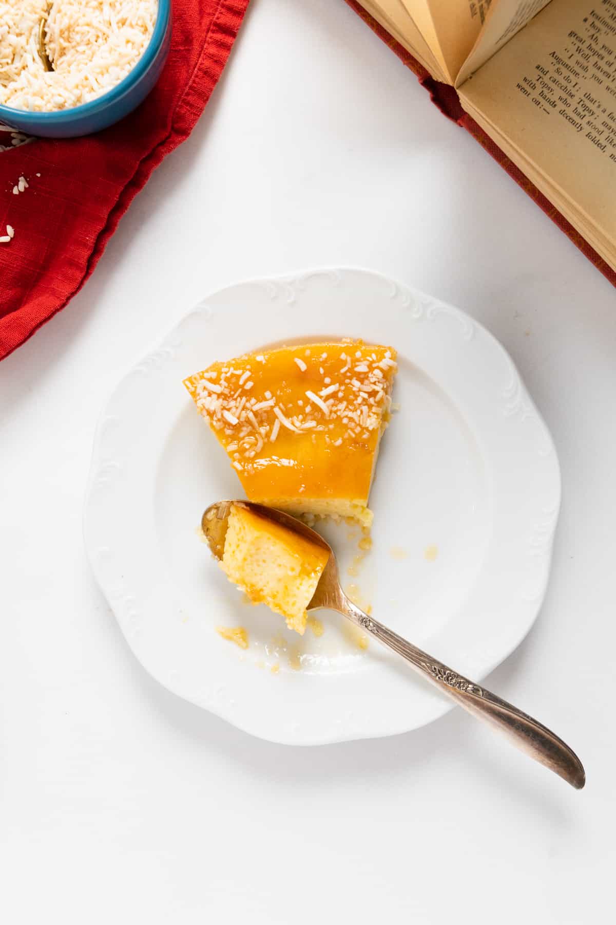 a slice of flan with a spoon and some flan in the spoon, an old book in the corner