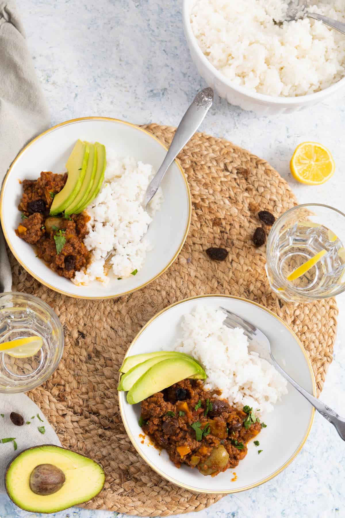 Ground beef served in white plate wit some white rice and 3 slices of avocados, there are 2 glasses of water with slices of lemon.
