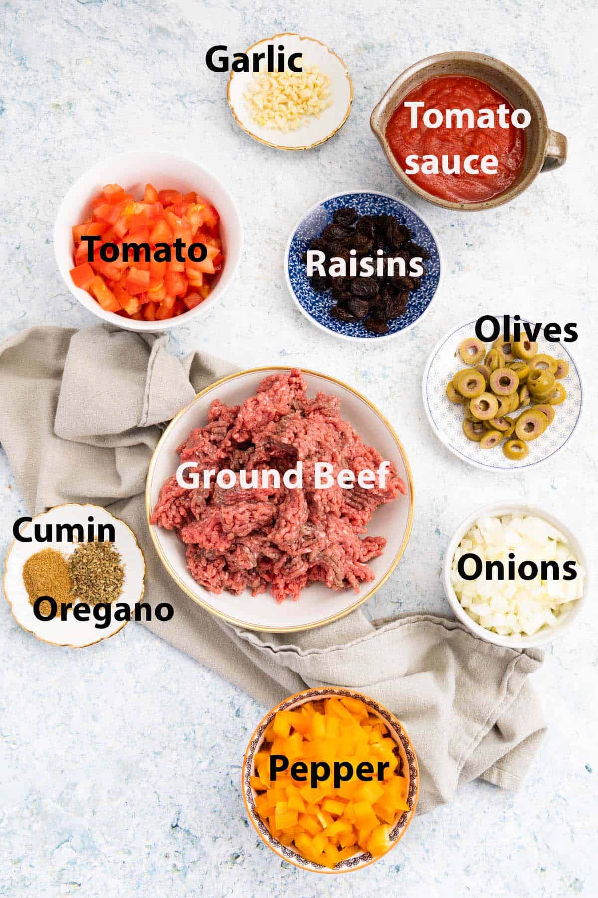 Ingredients in ramekins and small plates: garlic, tomato sauce, tomato, olives, ground beef, onions, pepper, cumin, oregano.