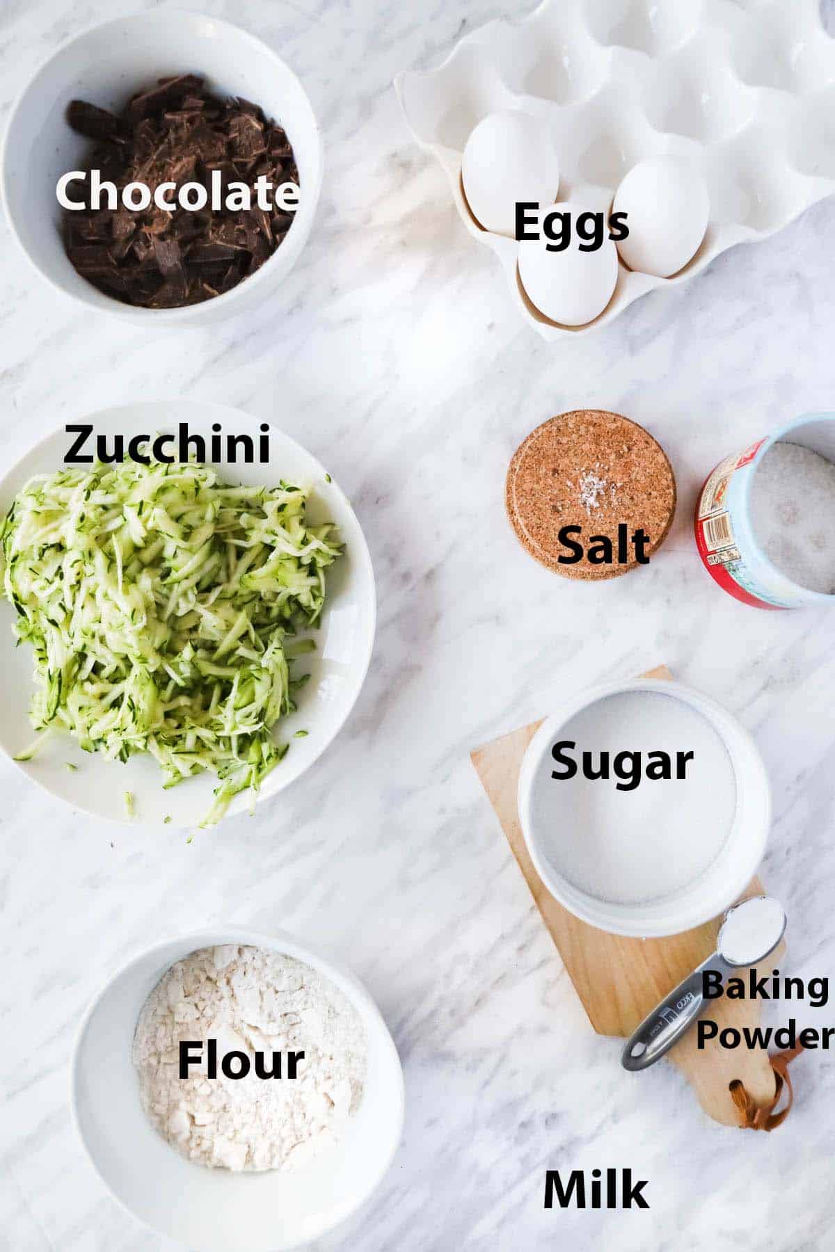 Labelled ingredients in bowls and ramekins: eggs, chocolate, shredded zucchini, salt, sugar, flour and baking powder.