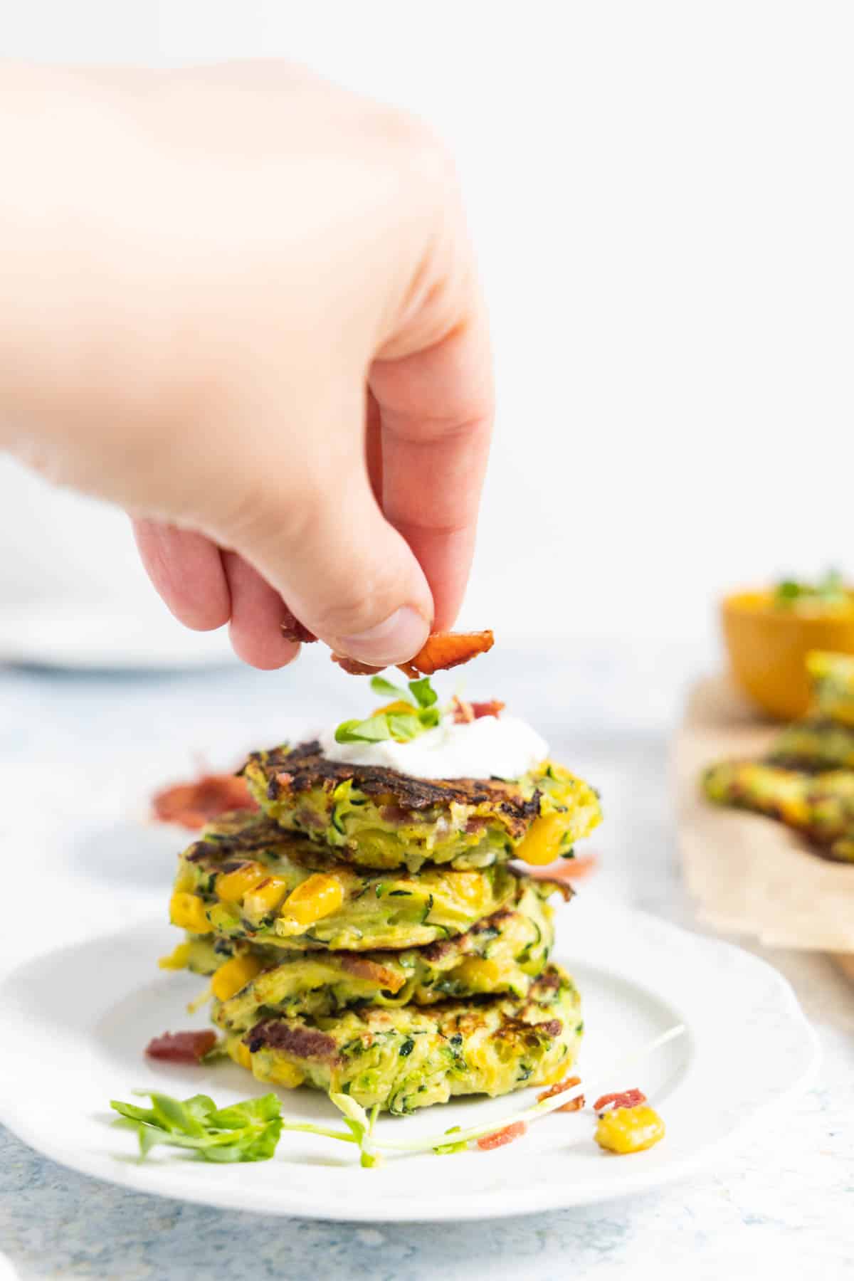 stack of 4 zucchini, corn and bacon fritters on a white plate with a dolop of sour cream on top and a hand sprinkling bacon. 