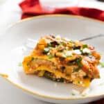 slice of lasagna in a white plate with gold rim and a fork in it and a glass of water in the background.