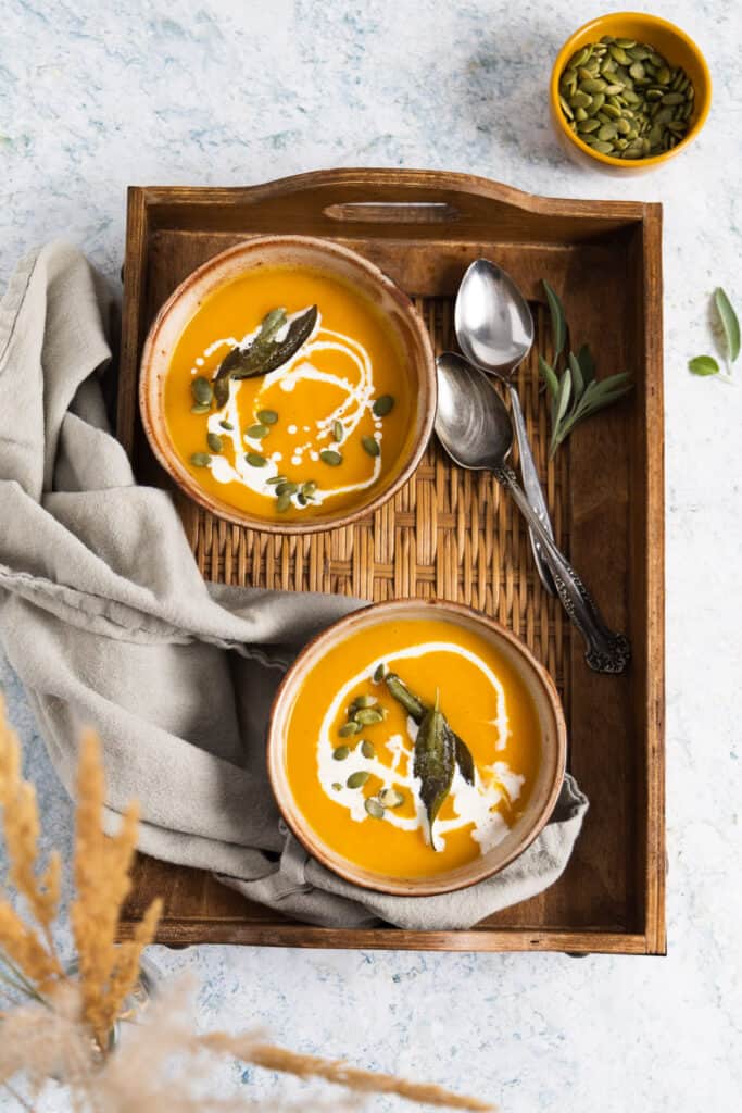 2 bowls of soup in a wooden tray with 2 spoons and a grey teatowel on the side.
