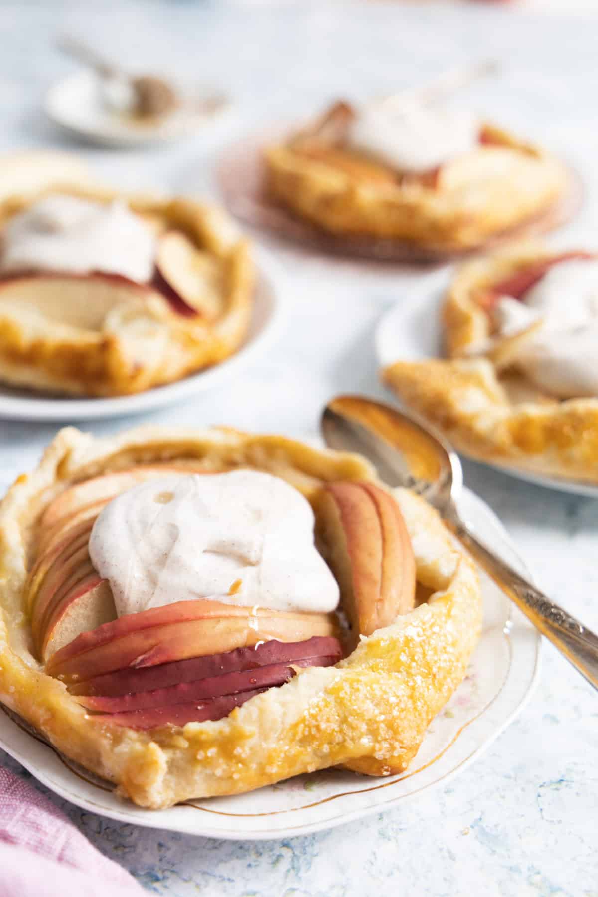 4 apple galettes with whipped cream on top, the first one has a spoon on a white plate. 
