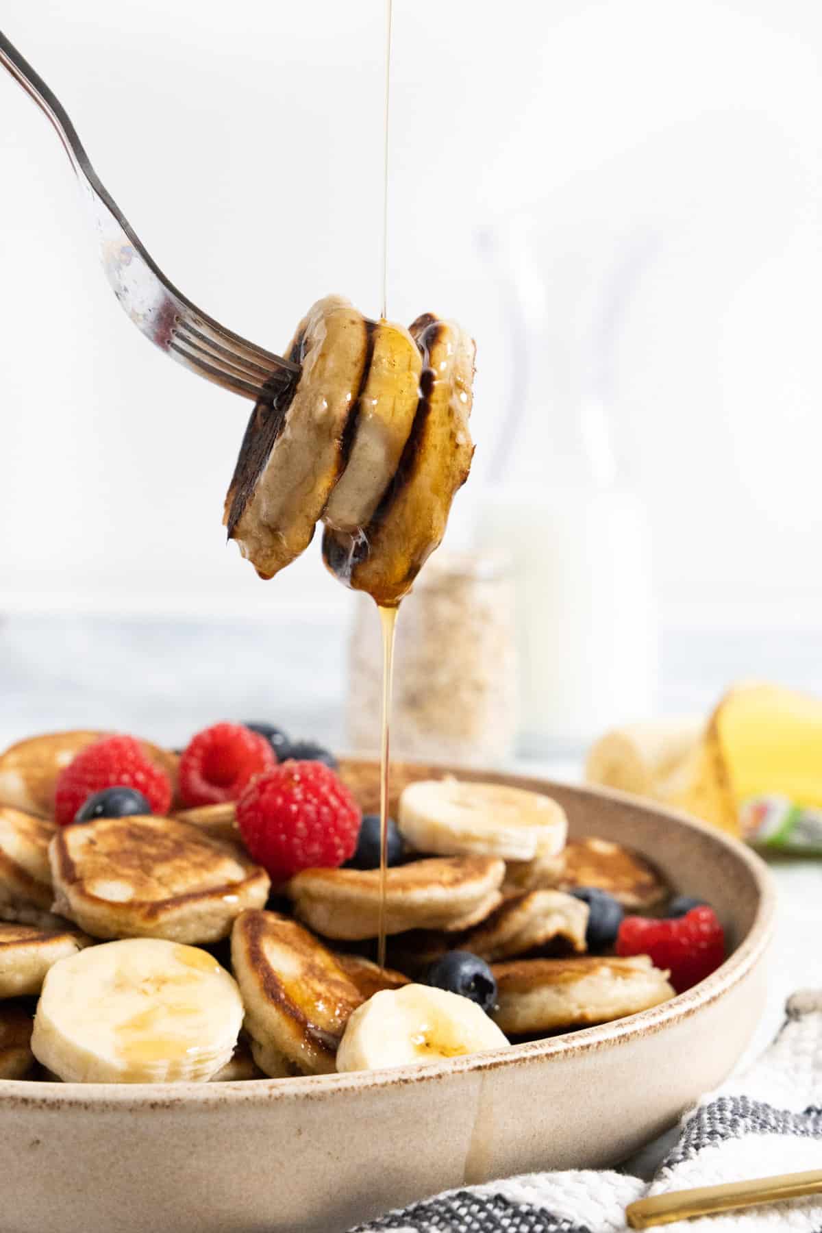 fork holding a slice of banana between 2 mini pancakes and a drizzle of maple syrup falling from it into a plate full of pancakes and fruit.