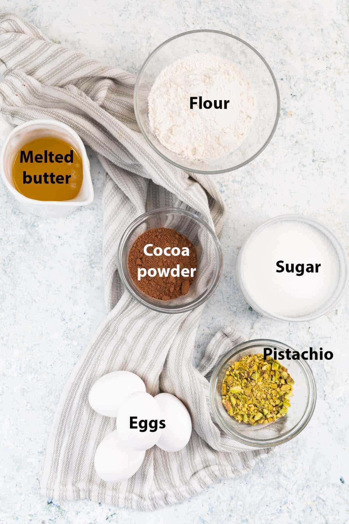 labelled ingredients in ramekins: flour, melted butter, sugar, cocoa powder, pistachio, eggs. 