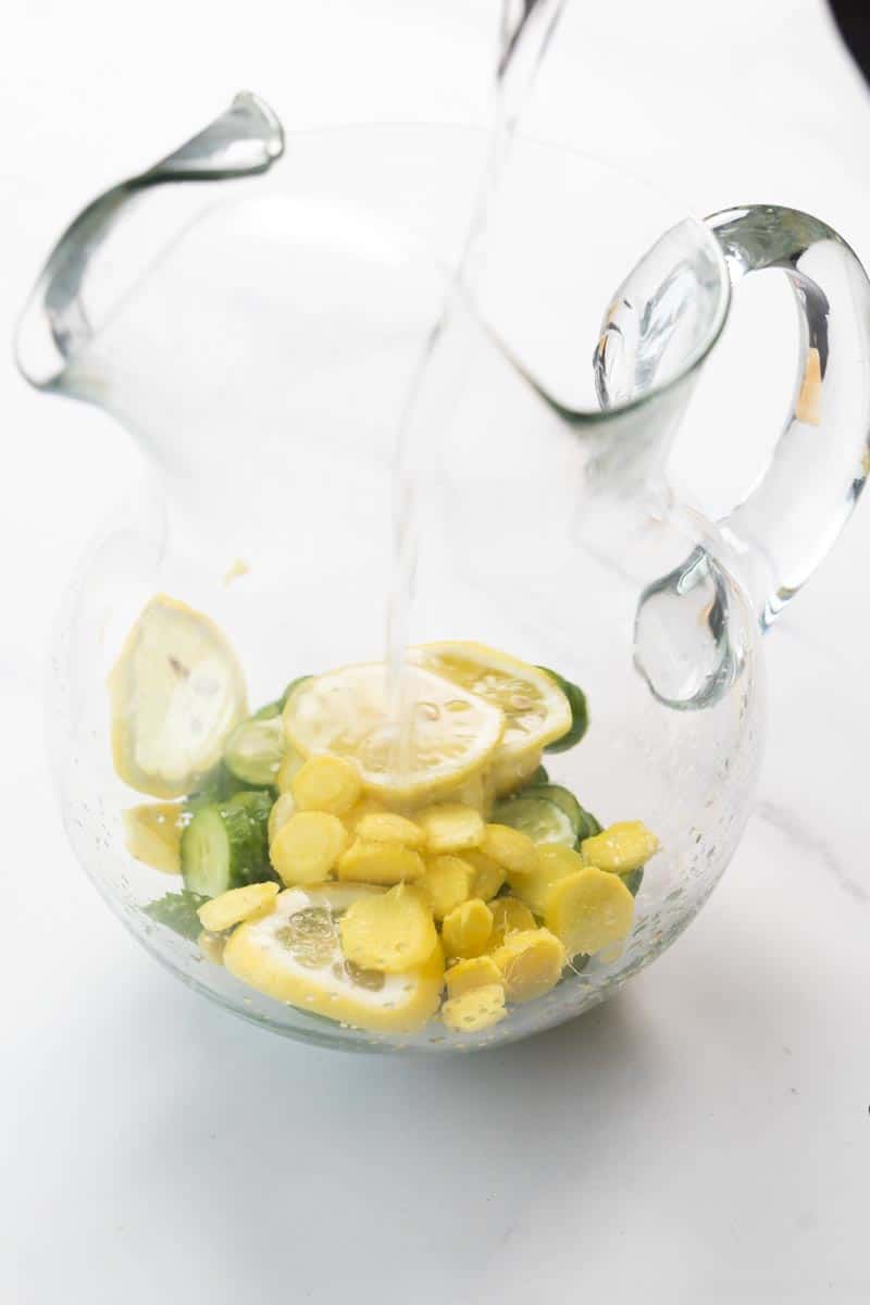 Sliced ginger, lemon and cucumber in a big transparent pitcher and water being poured. 