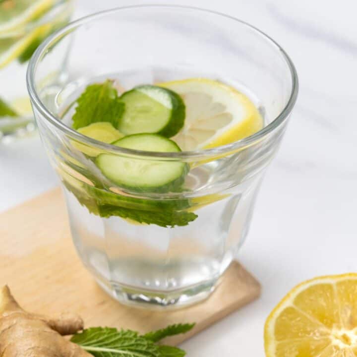 glass filled with water, 2 cucumber slices and a lemon slice are floating with mint leaves and half a lemon on the side.