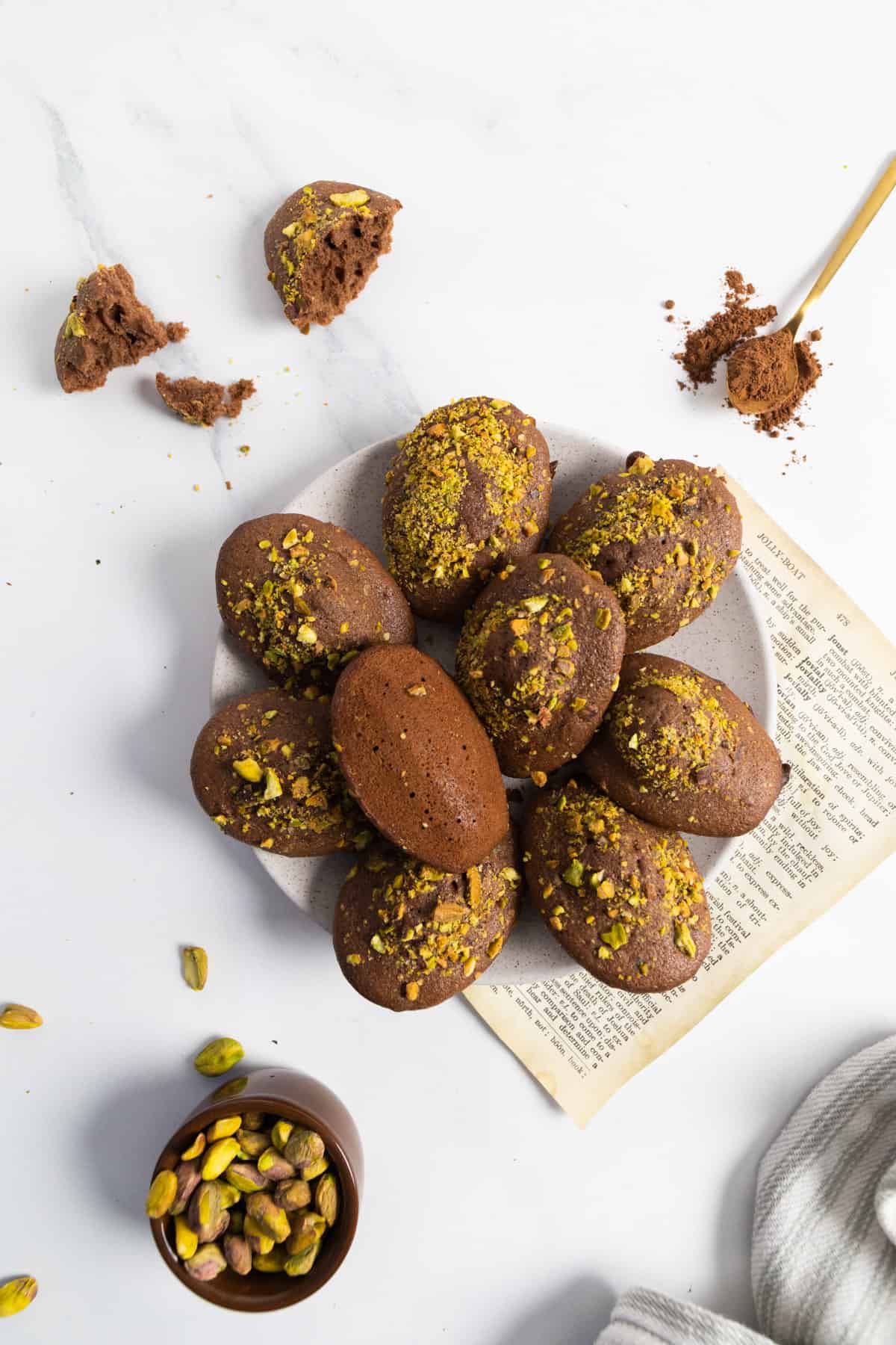 lots of chocolate and pistachio madeleines on a plate with a vintage piece of paper on the table, a ramekin on the side filled with pistachios. 