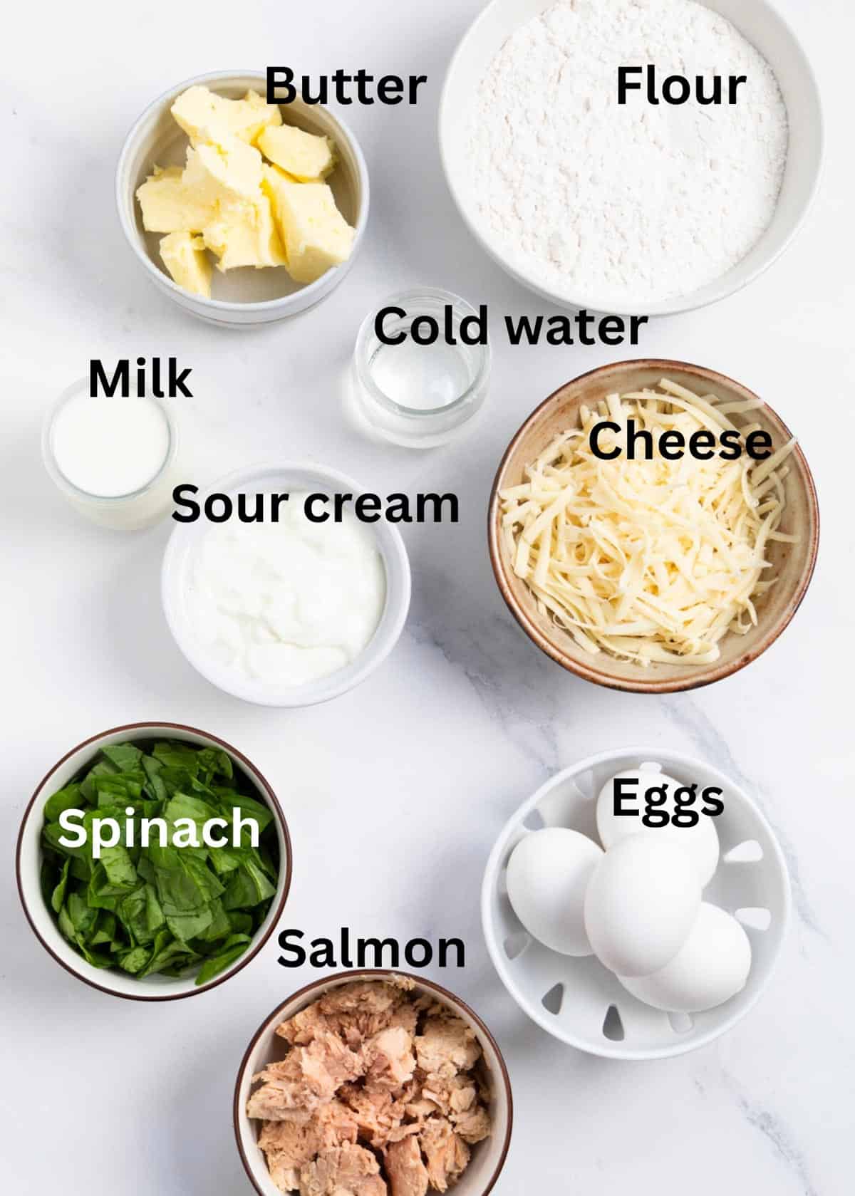 labelled ingredients on a marble background: butter, flour, cold water, cheese, milk, sour cream, spinach, salmon, eggs. 