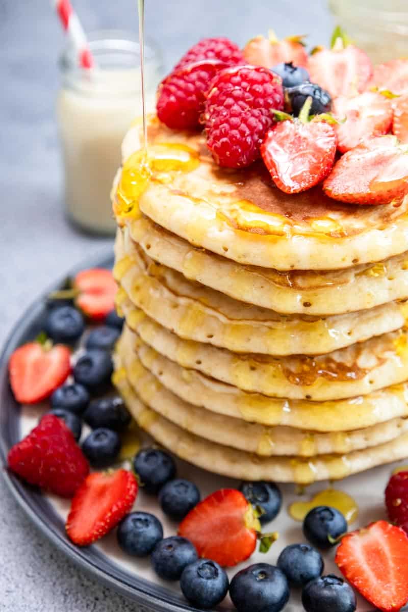 stack of 7 pacankes with strawberries, raspberries on top and in the plate with a drizzle of honey falling on the edge of the first pancake. 